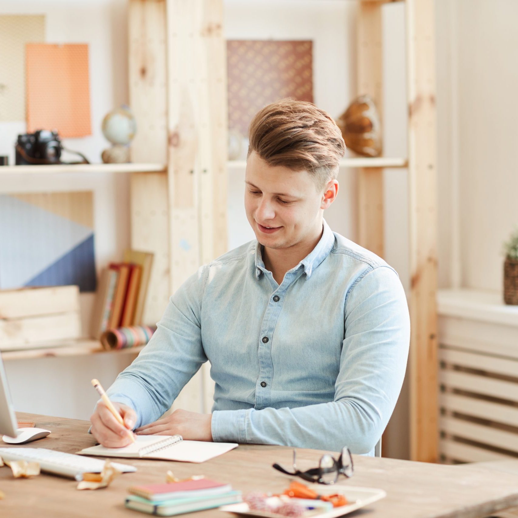 Content confident young creative manager in casual shirt sitting at desk with computer and making notes in diary while working in cozy office